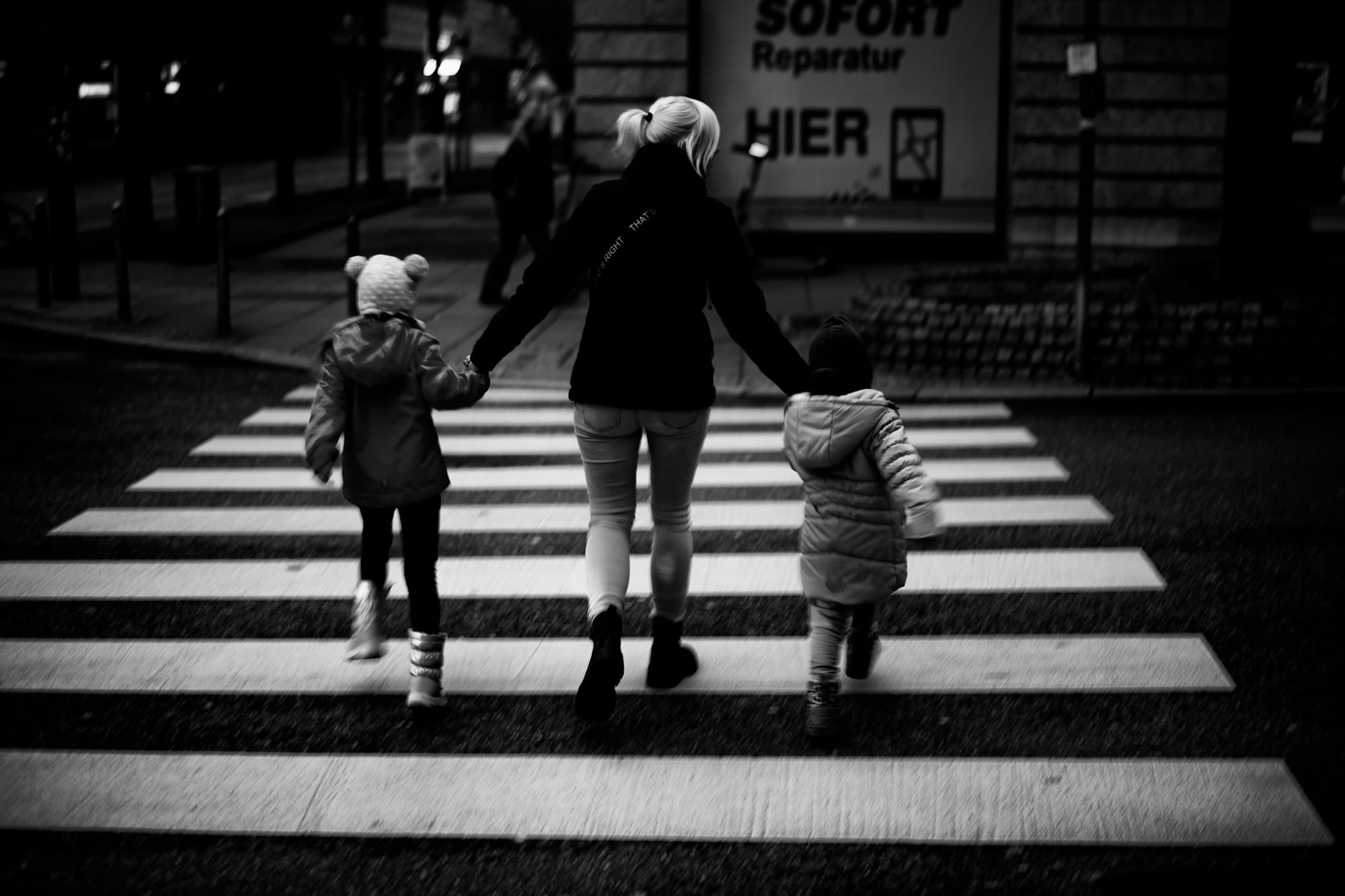 A mother and her two young children using a zebra crossing, from behind, in black and white.